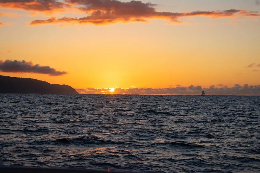 Sunset over the ocean with a distant sailboat and a silhouette of land against a vibrant orange and yellow sky.