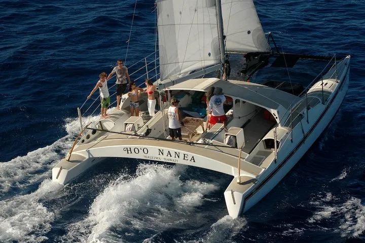 A white catamaran named HO'O NANEA with several people on board sails through deep blue ocean water.