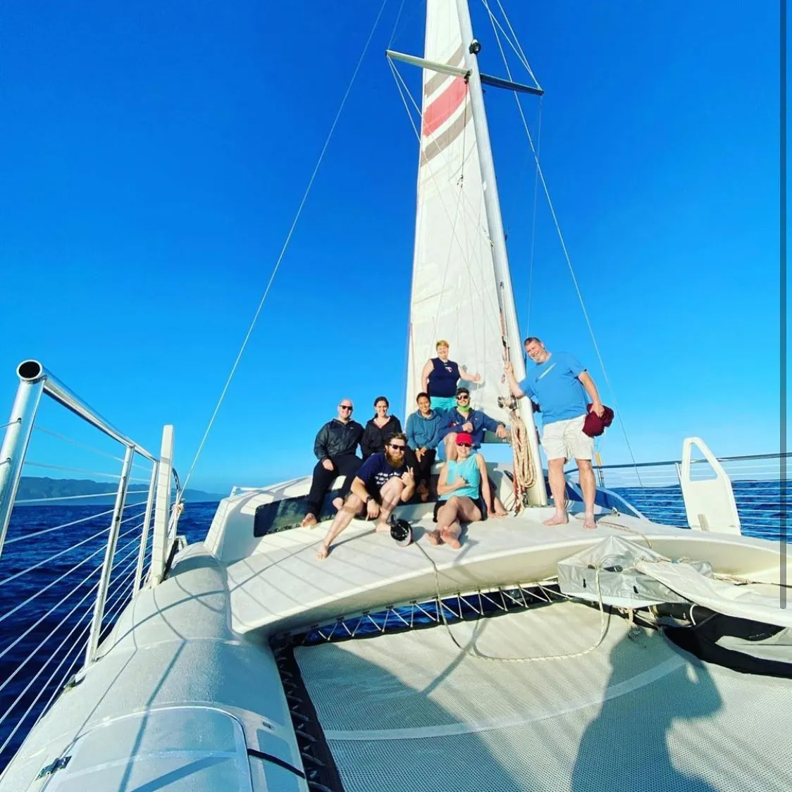 A group of eight people posing on the front deck of a large white catamaran sailing on clear blue water.