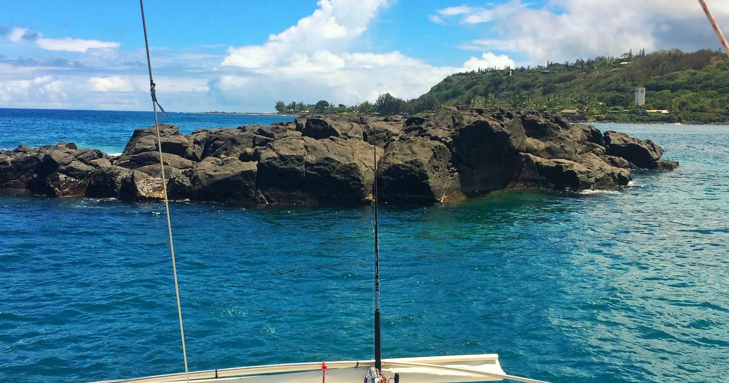 View from a boat of a large dark rock formation in clear blue water with a lush green coastline in the distance.