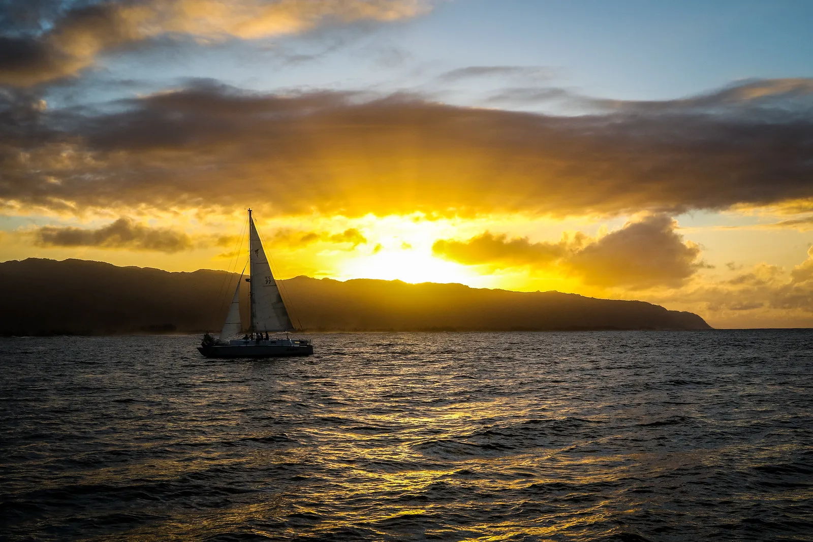 A sailboat on the ocean during a dramatic golden sunset with mountain silhouettes in the distance.