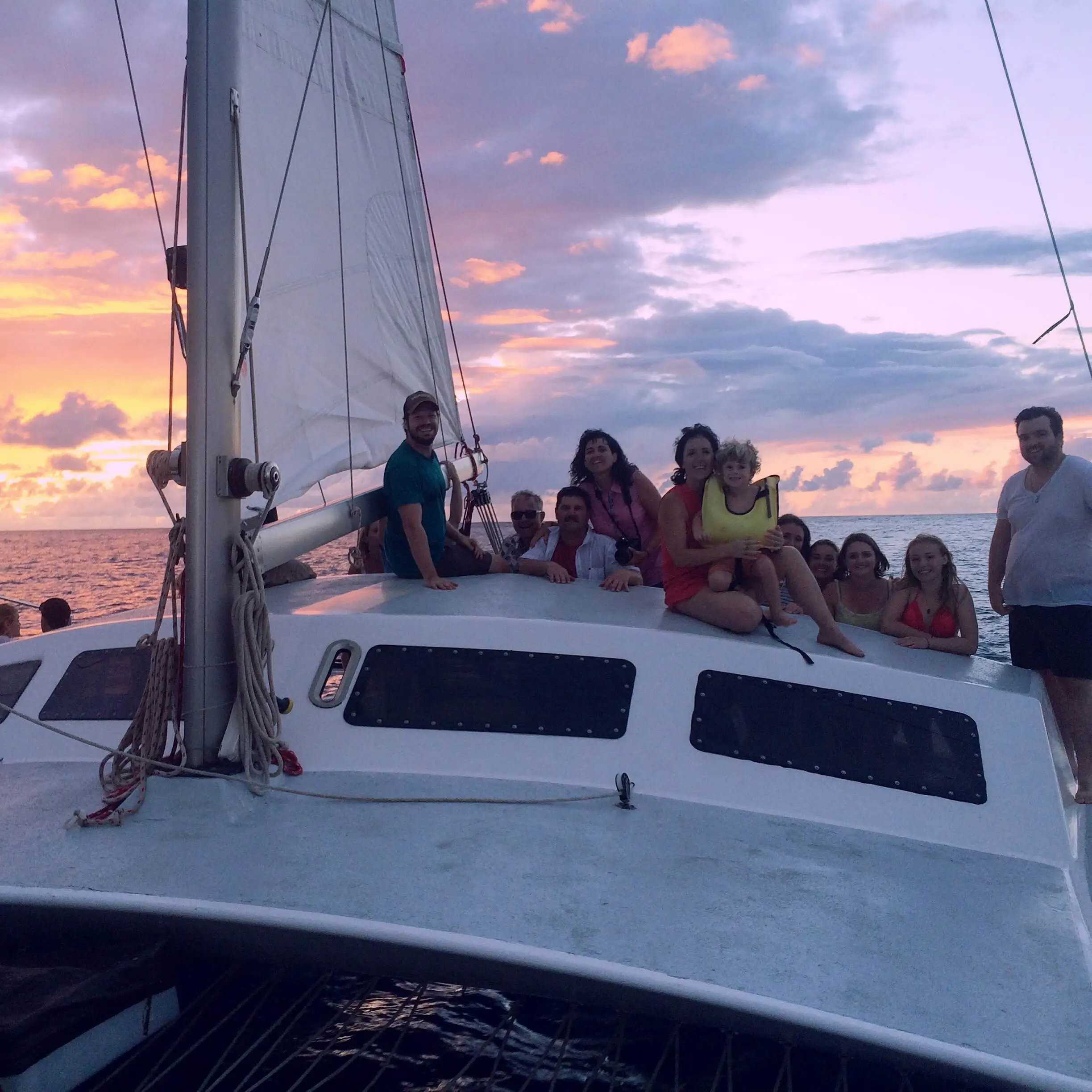 A group of people smiles on a catamaran deck during a vibrant sunset with pink and orange clouds.