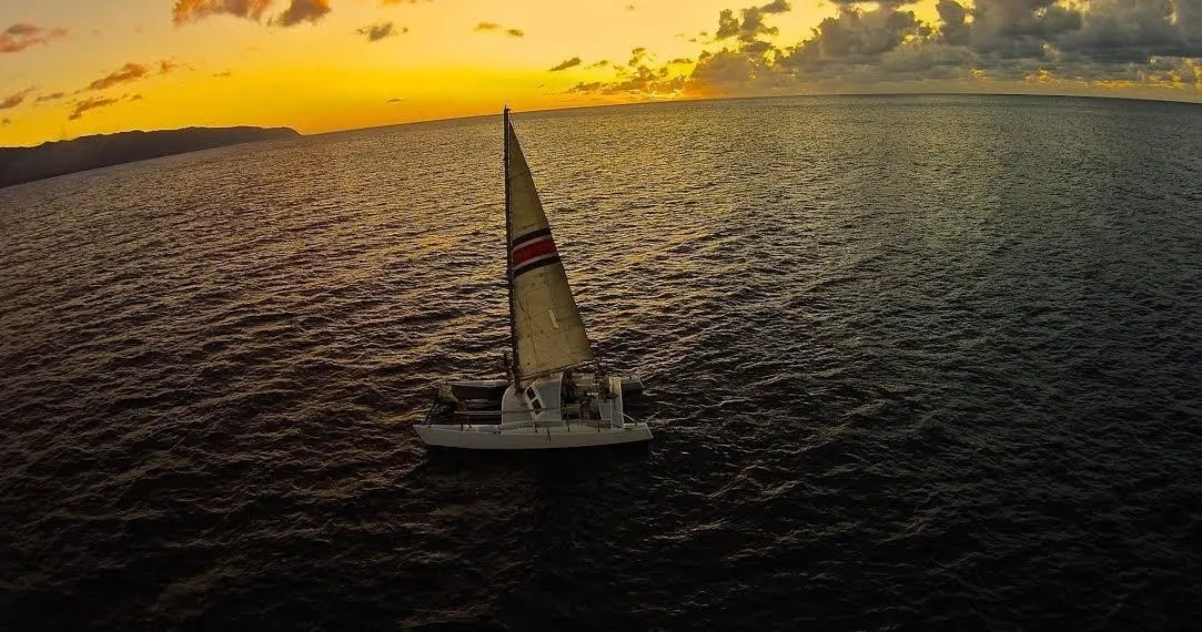 A white catamaran sailboat on a dark ocean under a vibrant orange and yellow sunset sky.