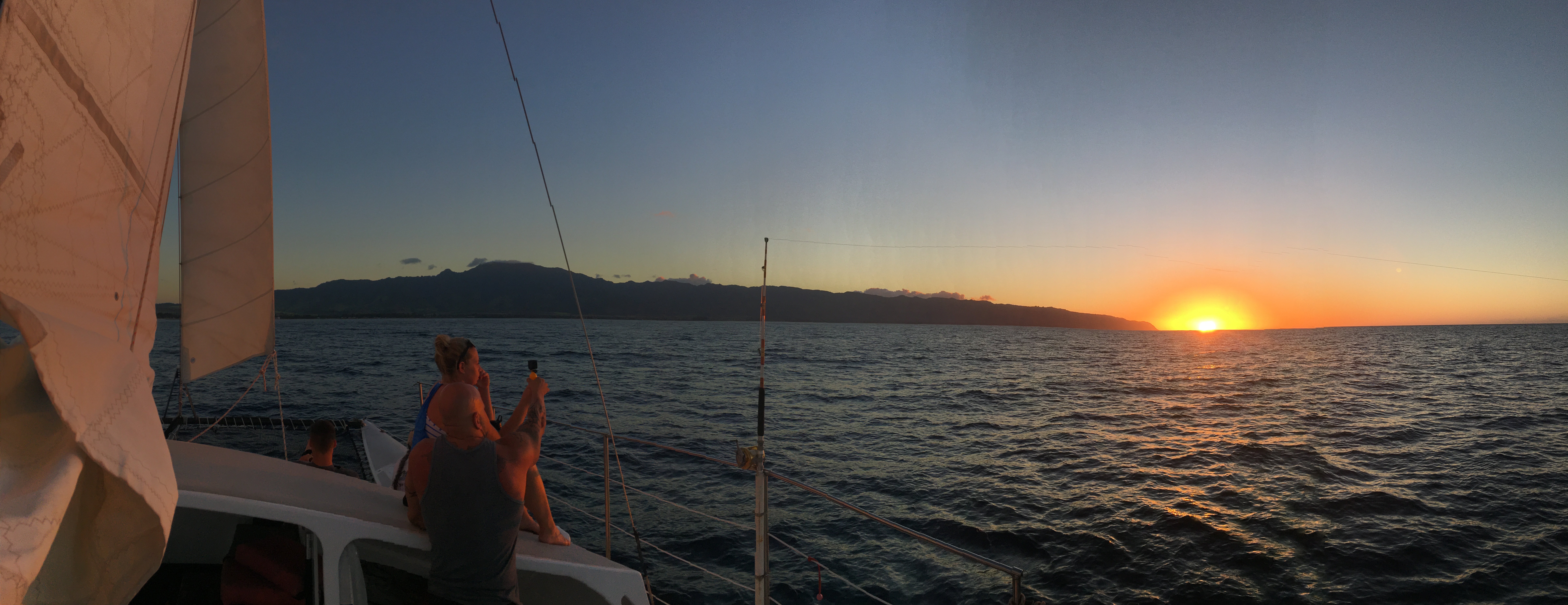 People on a sailboat watch a vibrant sunset over the ocean with dark mountains on the distant horizon.
