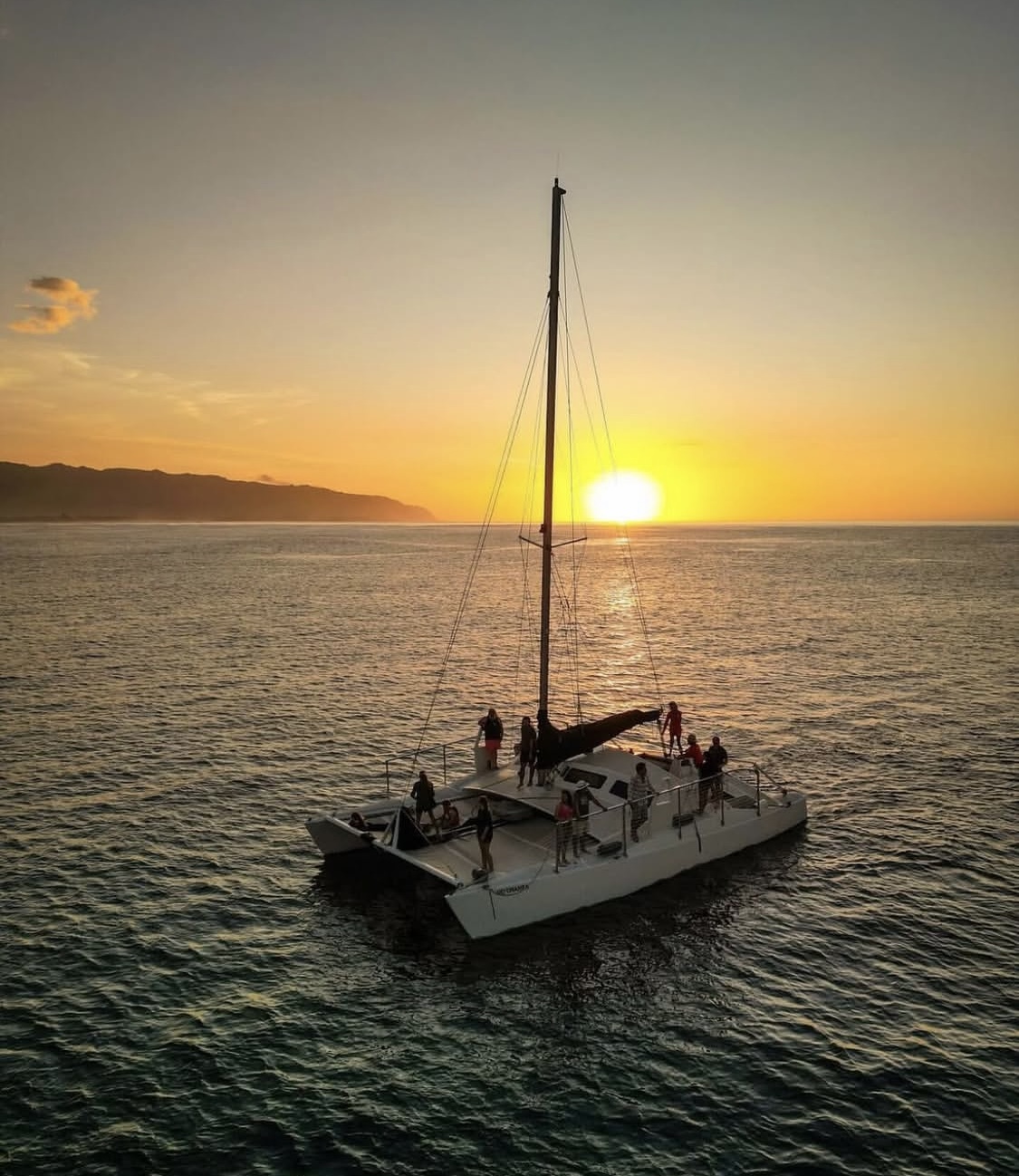 A catamaran carrying passengers sails on a calm ocean toward a bright golden sunset with mountains in the background.