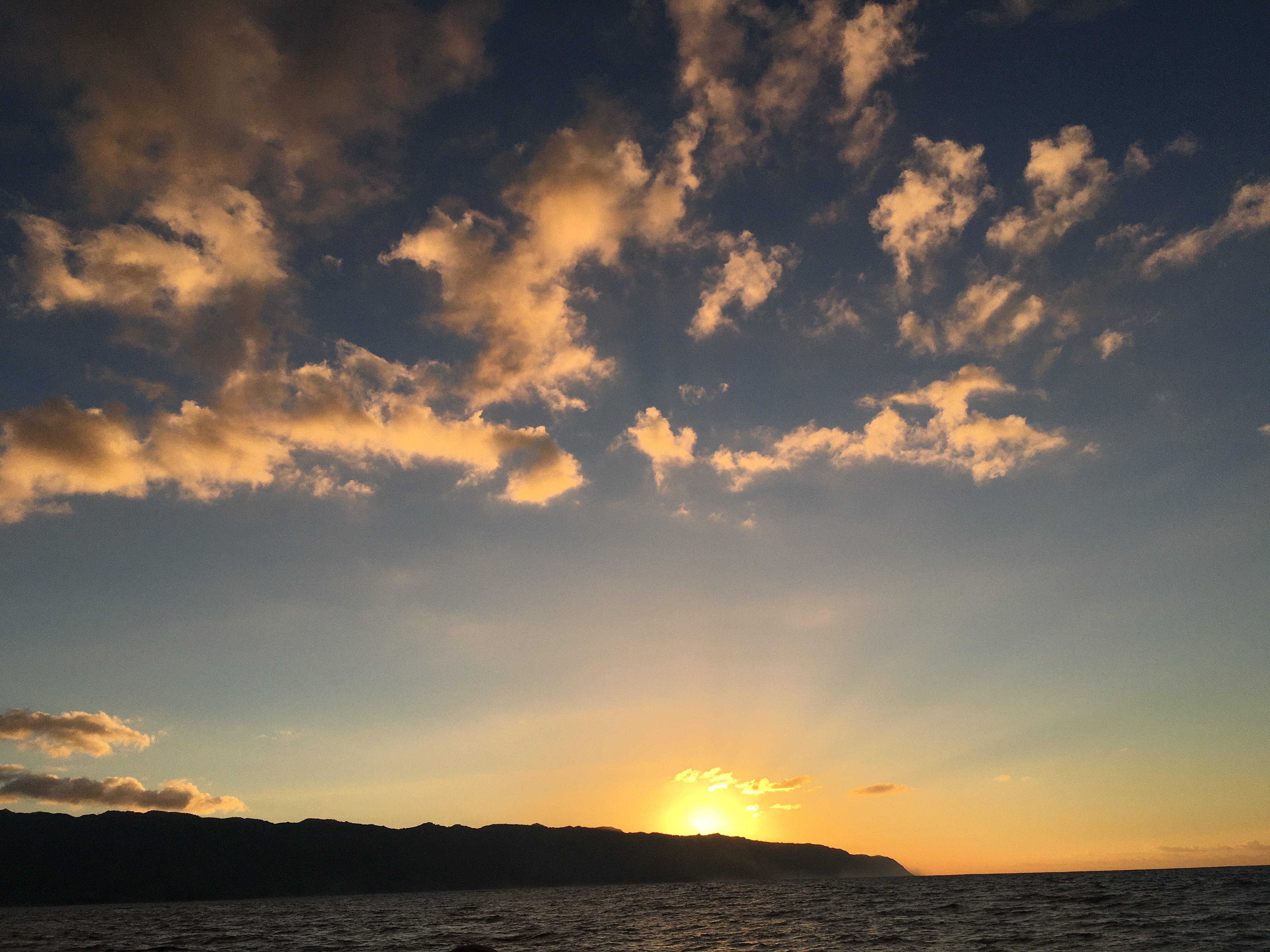 A golden sunset over silhouetted mountains and a calm ocean with scattered clouds catching the light.