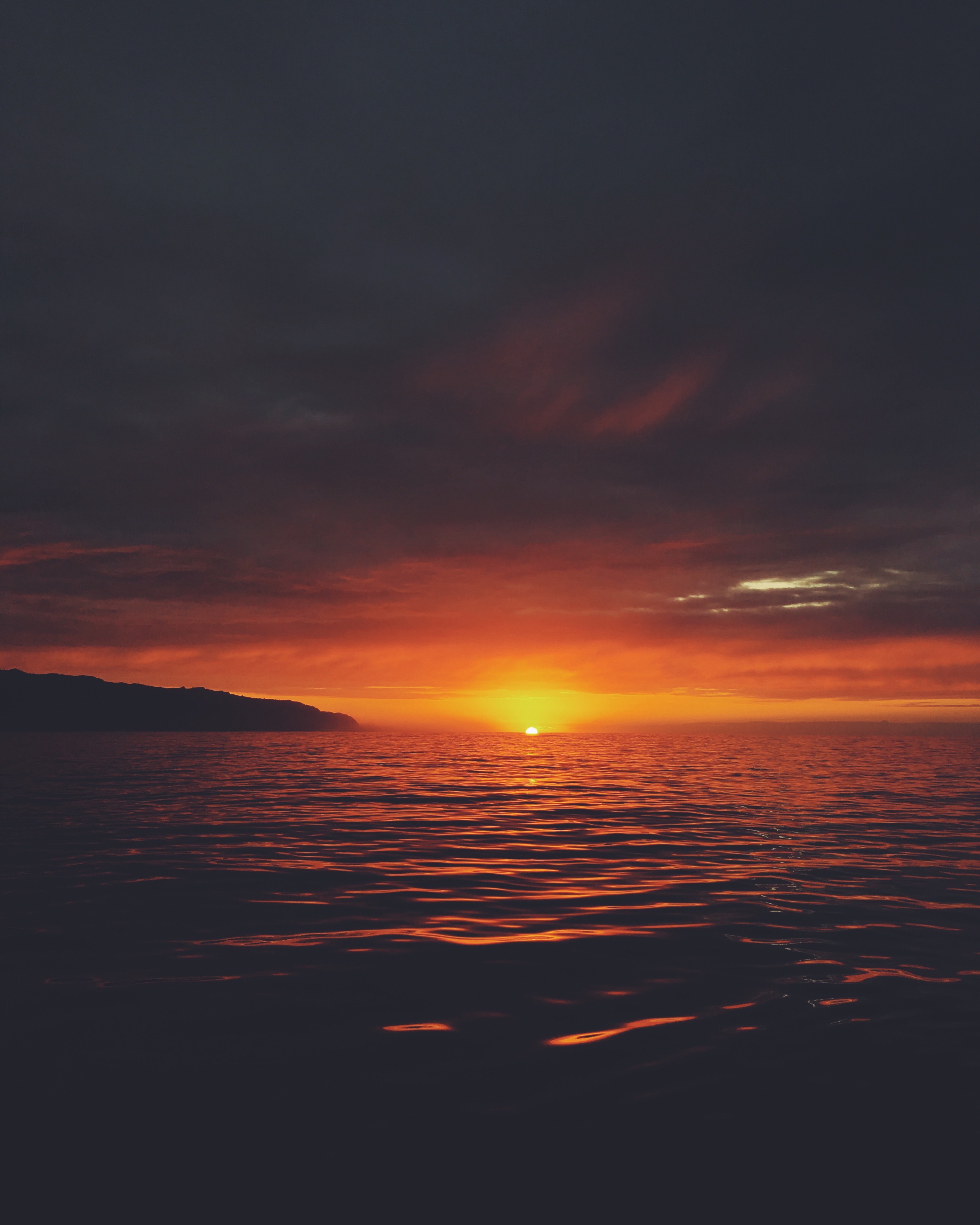 Sunset over a dark ocean with orange light on the horizon and a silhouette of land to the left.