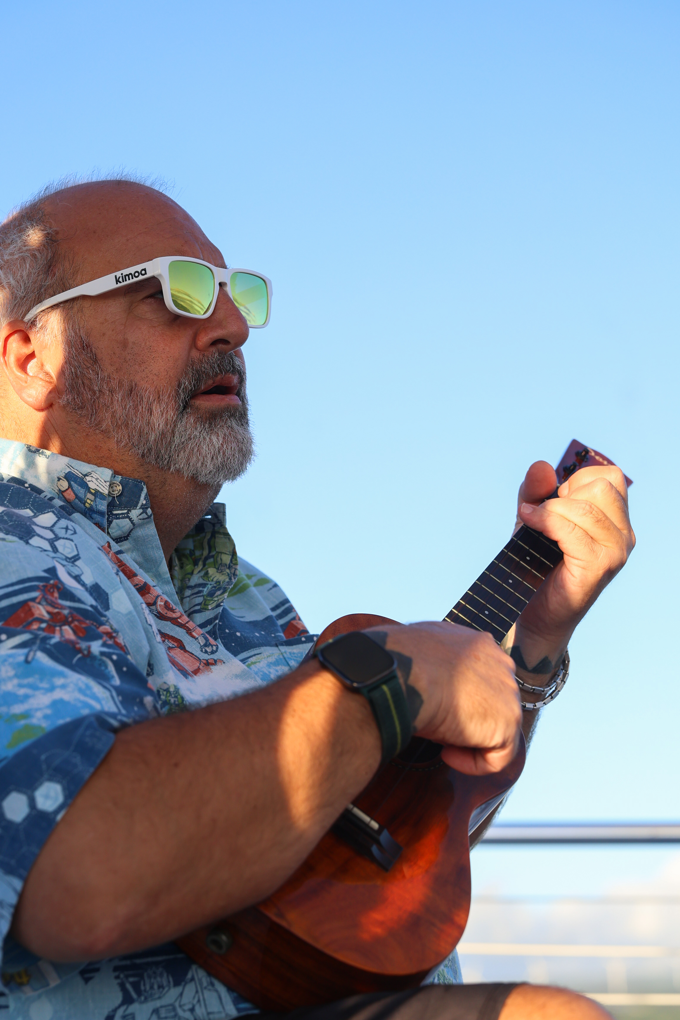 A man with a grey beard and green-lensed white sunglasses plays a ukulele against a clear blue sky.