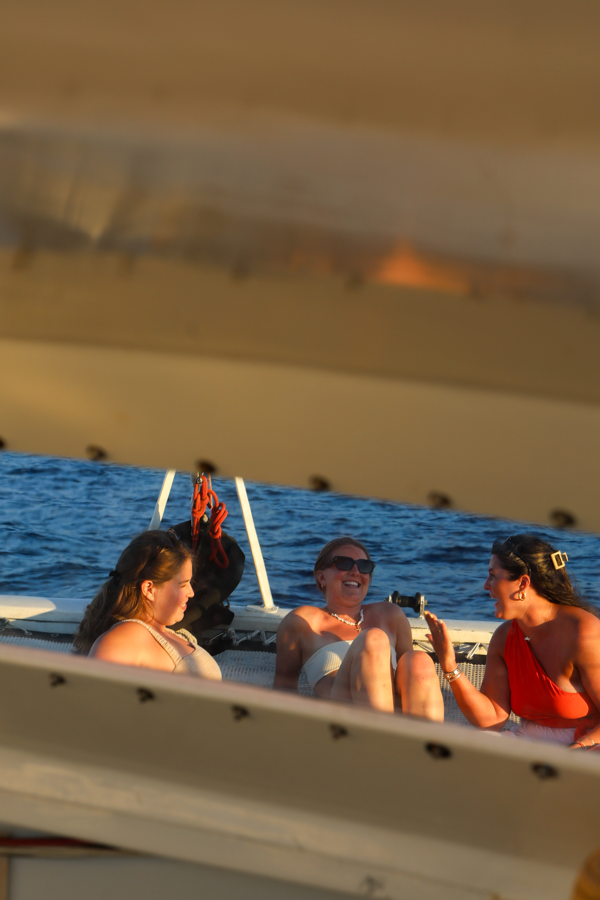 Three women laughing together while lounging on a boat deck at sea, framed by architectural elements.