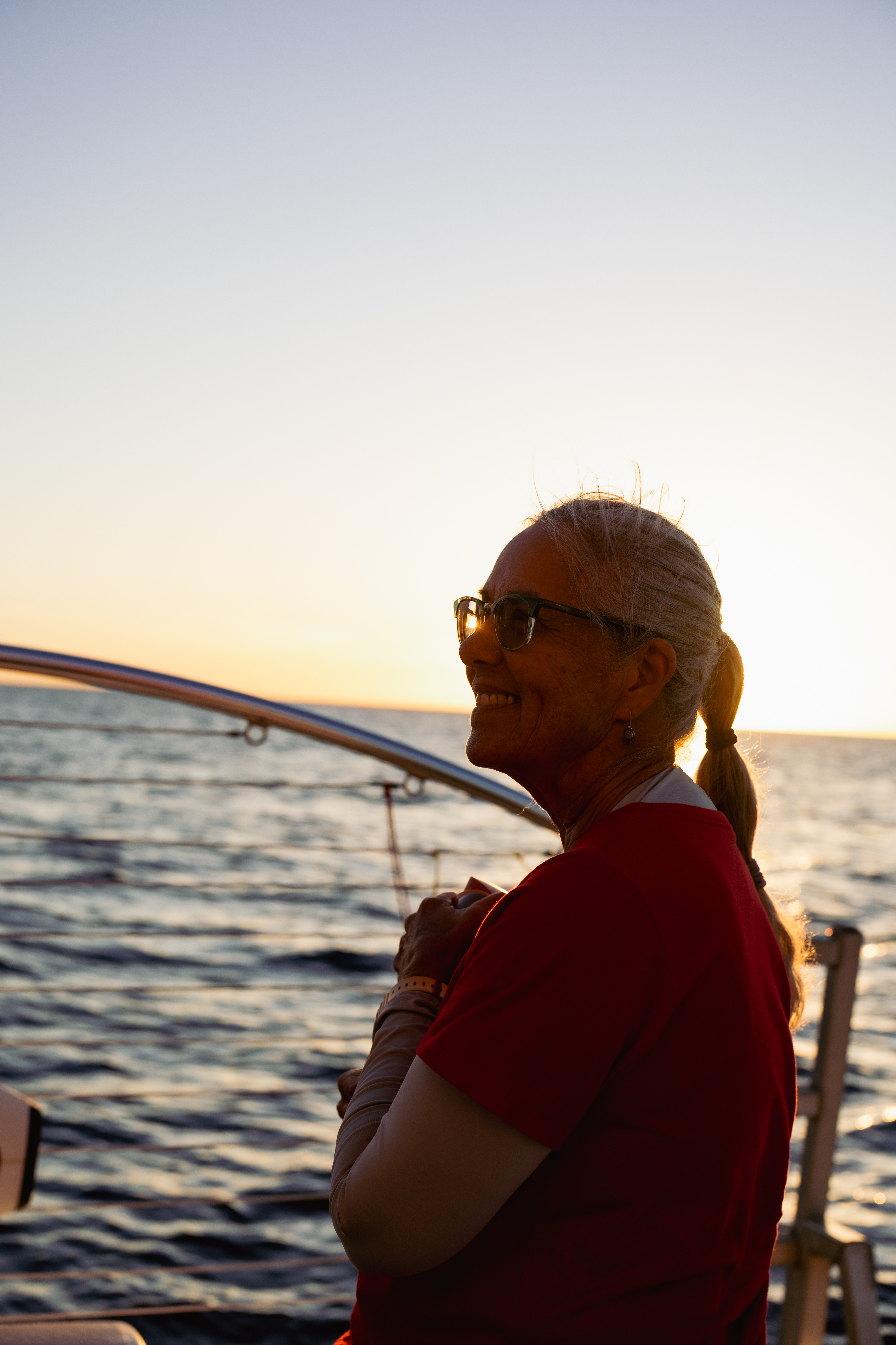 A smiling older woman with grey hair in profile on a boat at sunset, looking out at the ocean.