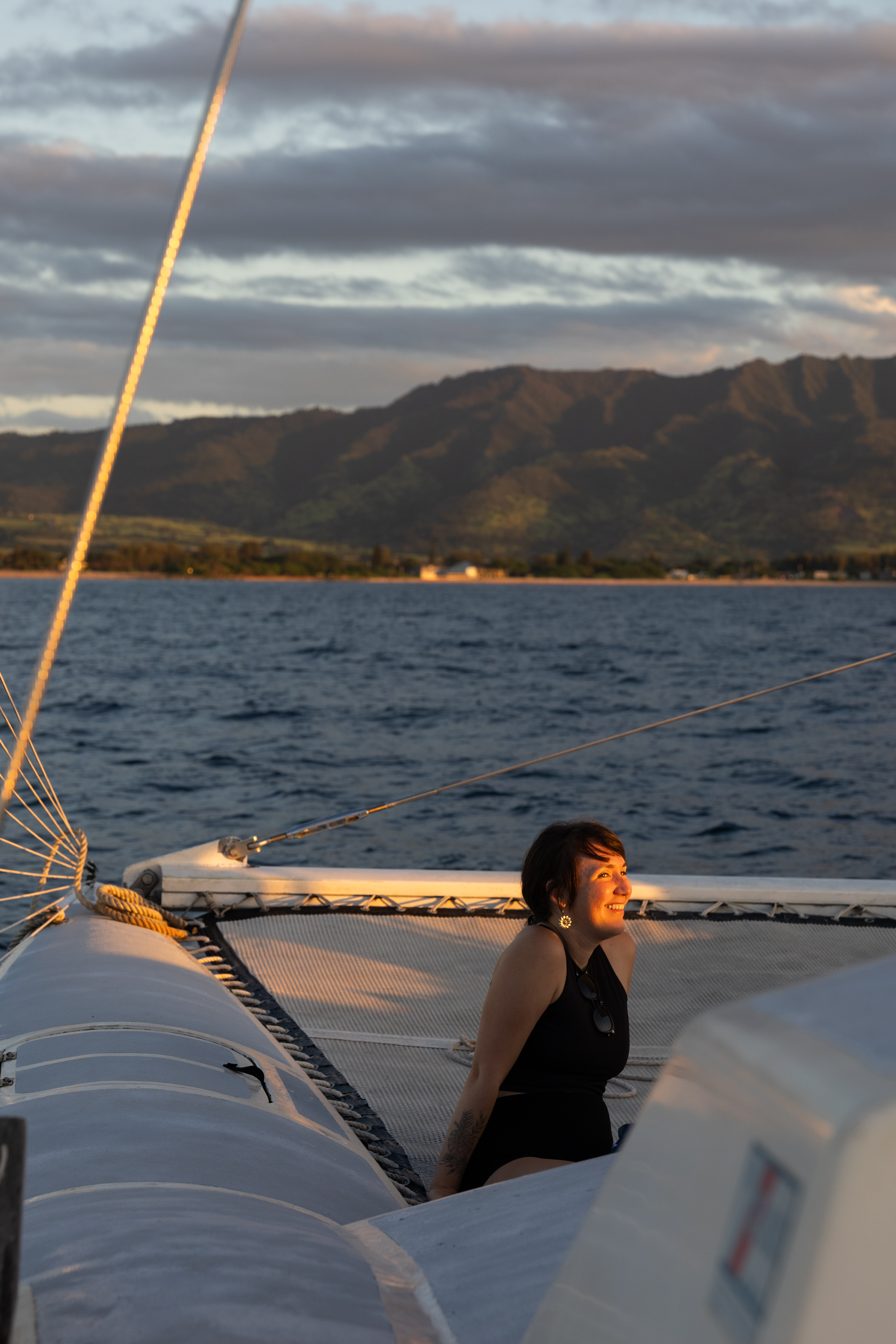 A smiling woman sits on a catamaran netting at sunset, with dark mountains and the ocean in the background.