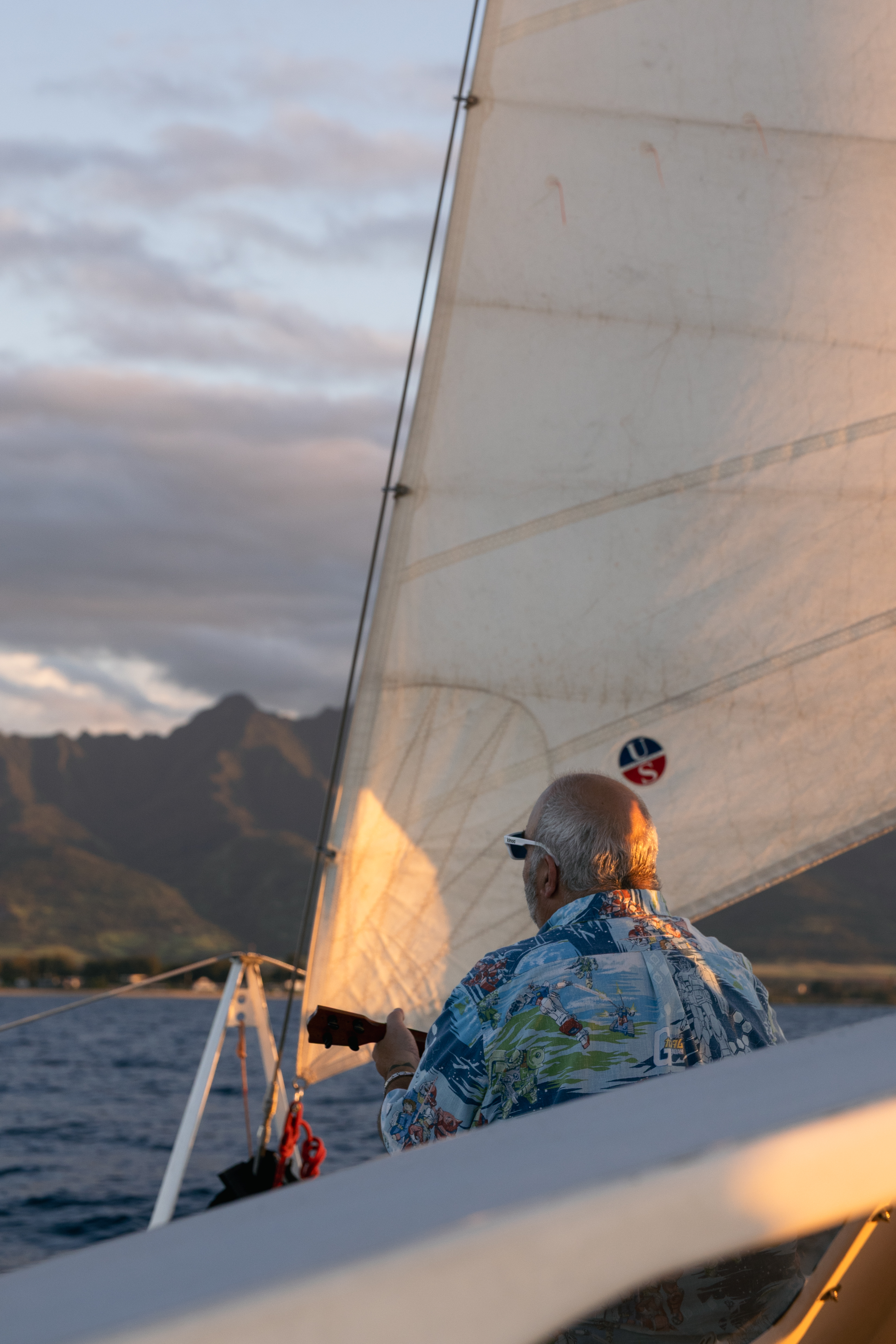 Older man in a patterned shirt plays a ukulele on a sailboat at sunset near mountains.