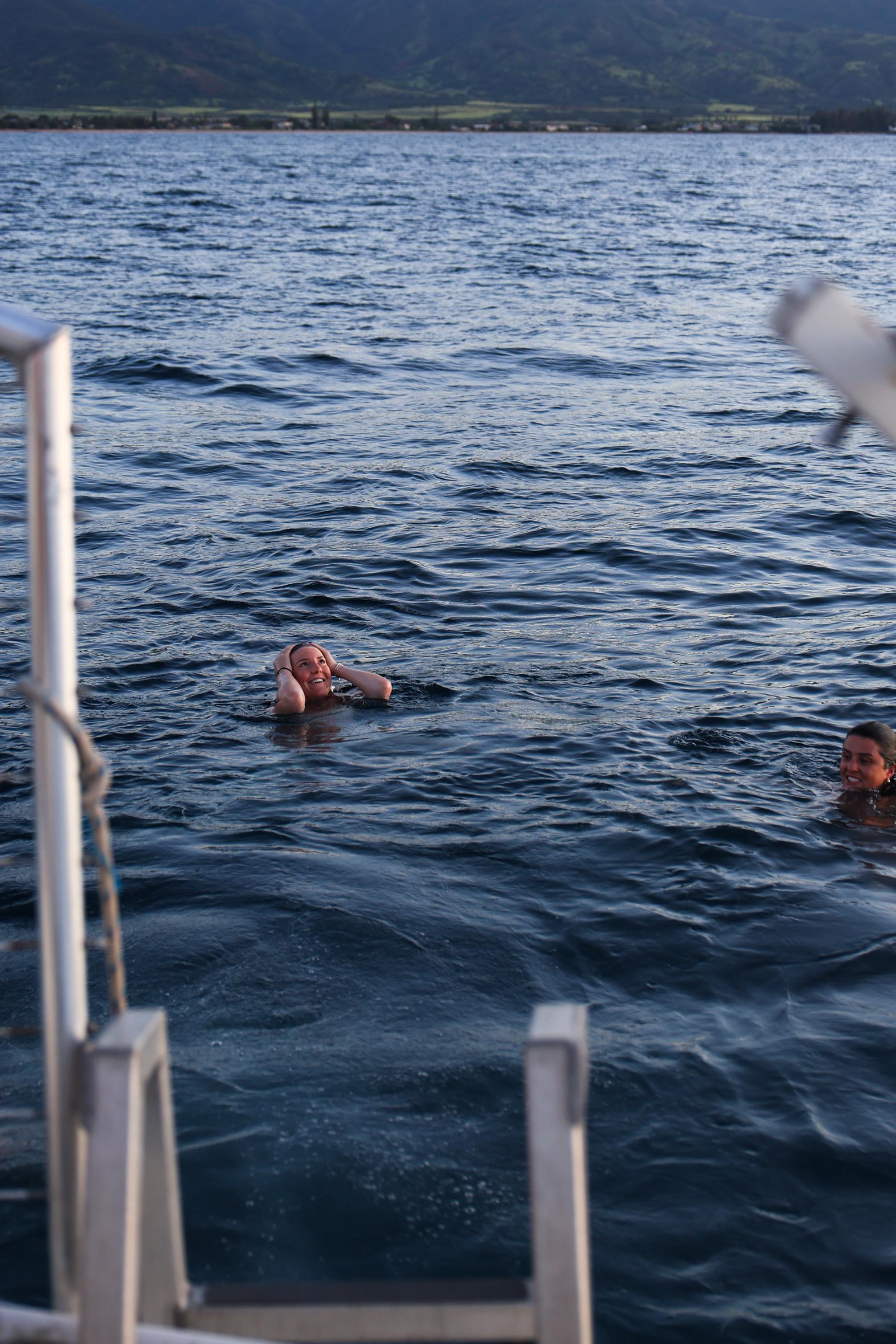 Two women swimming in dark blue ocean water near a boat ladder with hazy mountains in the distance.