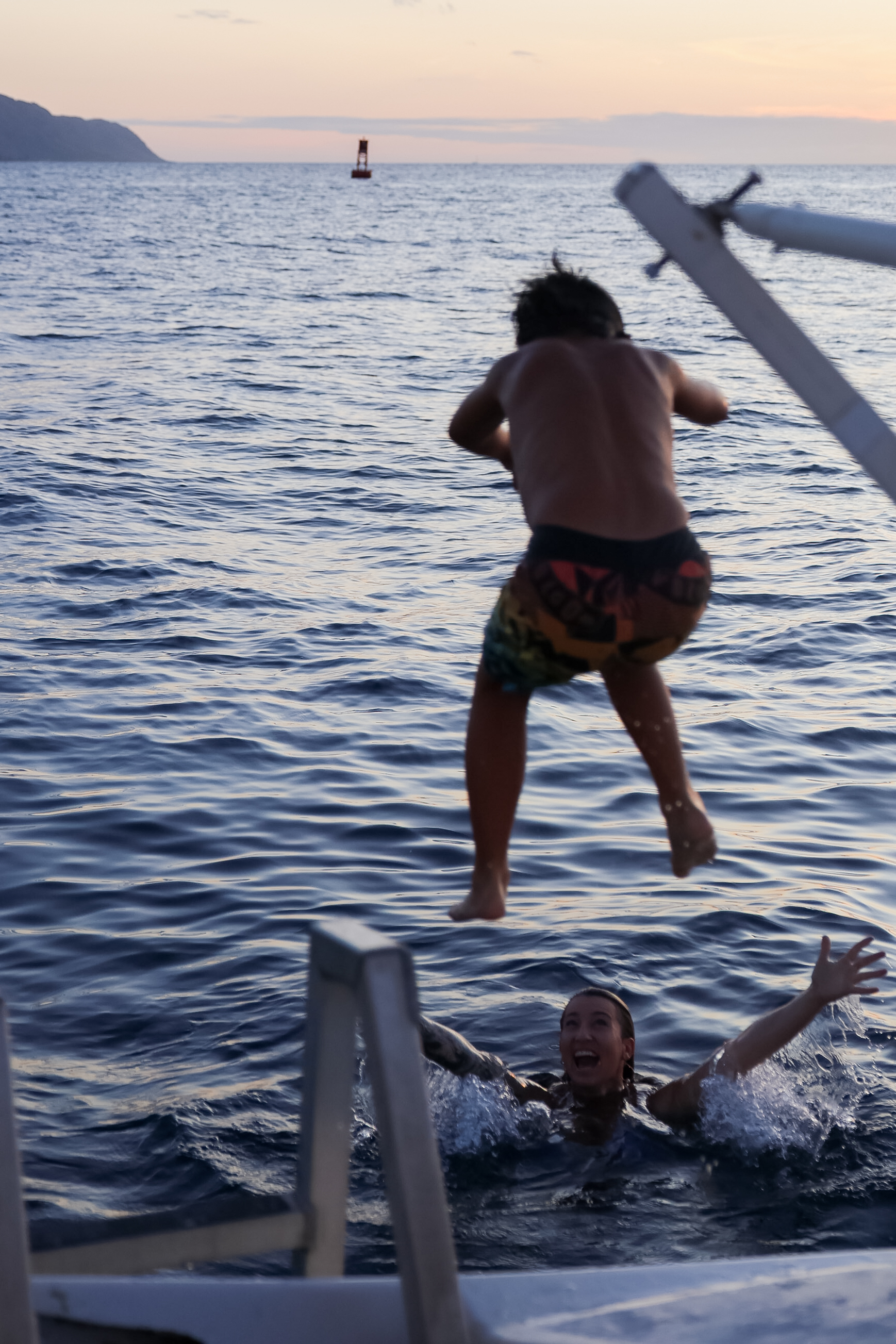 A person jumps off a boat into the ocean as another person in the water below watches with arms open during sunset.